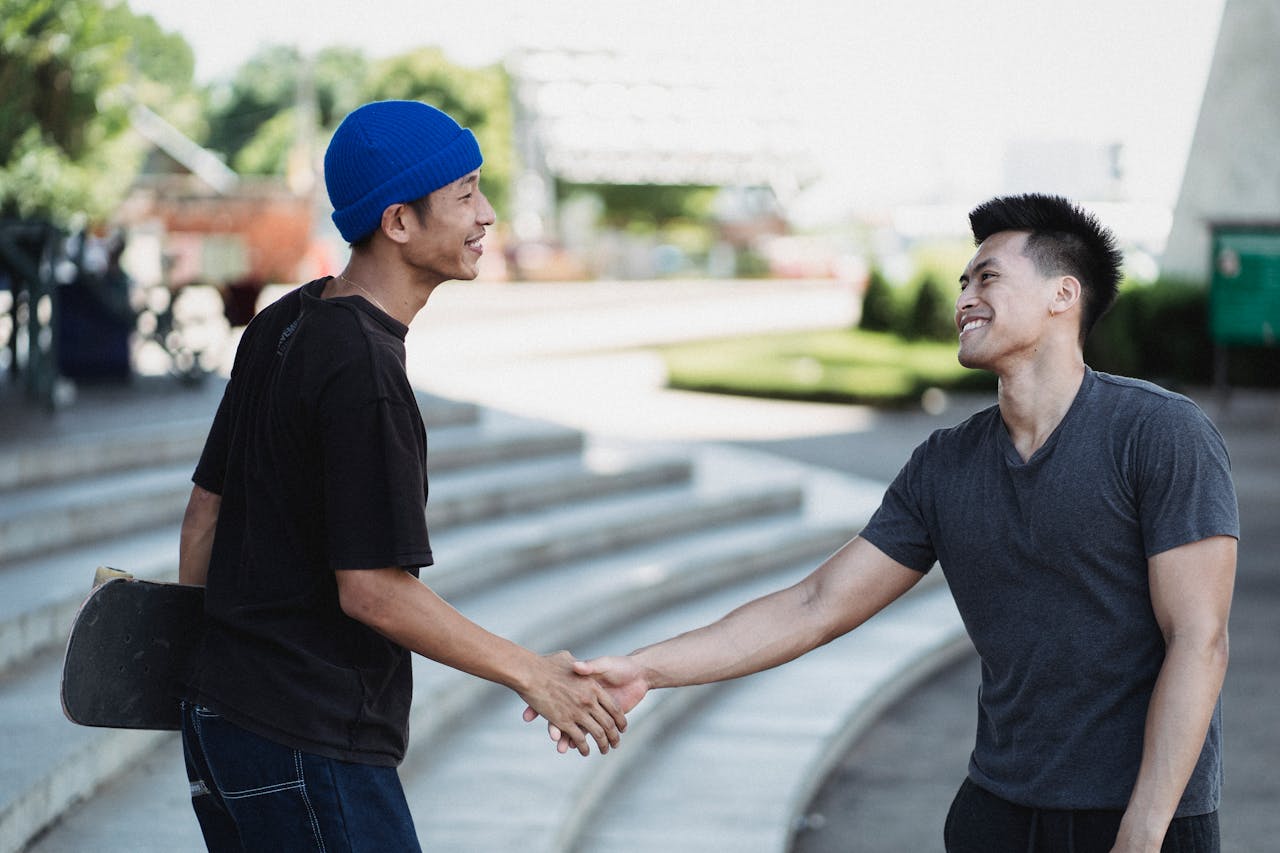 Cheerful young Asian men in street wear shaking hands and greeting each other with friendly smile